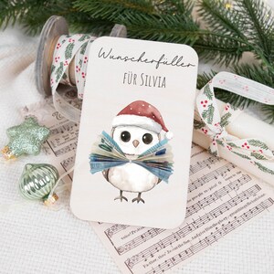 May include: A white card with a cartoon owl wearing a Santa hat and holding a fan of money. The card says "Wunscherf&uuml;ller f&uuml;r Silvia" which translates to "Wish Fulfillment for Silvia".
