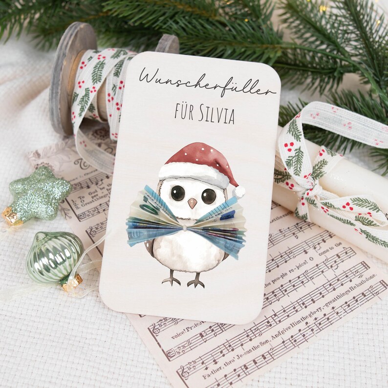May include: A white card with a cartoon owl wearing a Santa hat and holding a fan of money. The card says "Wunscherf&uuml;ller f&uuml;r Silvia" which translates to "Wish Fulfillment for Silvia".