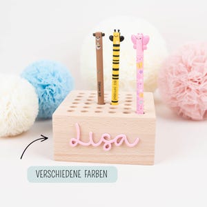 May include: A light wooden pen holder with a pink name, "Lisa", and three decorative pens. The pens have animal-themed designs: a bear, a bee, and a butterfly. The text "VERSCHIEDENE FARBEN" is at the bottom.