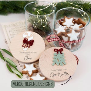 Puede incluir: Posavasos de madera con diseños festivos, incluyendo un bastón de caramelo y un árbol de Navidad, junto a galletas en forma de estrella en un tarro de cristal. Los posavasos tienen texto en alemán. La imagen también incluye vegetación y luces de hadas.