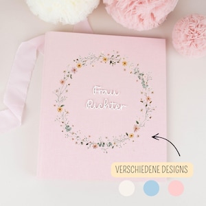 Può includere: Un libro rosa chiaro con un'illustrazione di una corona floreale al centro. Il testo "Frau Richter" è stampato in bianco al centro della corona. Una freccia nera punta verso il testo e un'etichetta recita "VERSCHIEDENE DESIGNS" con tre cerchi.