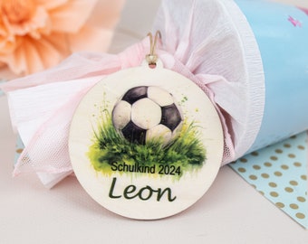 Pode incluir: Uma etiqueta redonda de madeira com uma ilustração de aquarela de uma bola de futebol em grama verde. A etiqueta tem o texto "Schulkind 2024" e o nome "Leon" impresso nela.