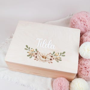 Può includere: Una scatola di legno con una finitura in legno chiaro e un design floreale. La scatola ha un'iscrizione bianca che dice "Tilola".