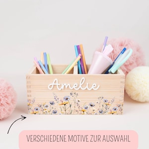 Op de afbeelding: Een lichtgekleurde houten kist met de naam "Amelie" in witte letters. De kist is versierd met een bloemenontwerp en bevat diverse pennen en potloden. Een roze beker en decoratieve ballen staan op de achtergrond.