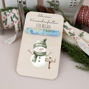 May include: A wooden card with a snowman illustration and a rolled up bill of money. The card says "Kleiner Wunscheref&uuml;ller f&uuml;r MILAN" which translates to "Small wish fulfillment for MILAN".