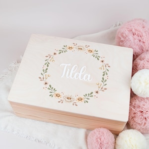 Può includere: Una scatola di legno chiaro con un design a corona di fiori e il nome "Tilda" scritto in lettere bianche. La scatola è circondata da pompon  rosa e bianchi.