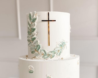 Op de afbeelding: Een witte bruidstaart met twee lagen, versierd met groene en gouden geverfde bladeren en een gouden kruis bovenop.