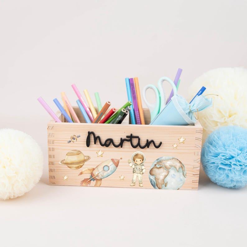 May include: A rectangular wooden pencil holder featuring the name Martin in black script. The front is decorated with watercolour illustrations of a planet, a rocket ship, an astronaut, and stars. It is filled with various coloured pencils and scissors.
