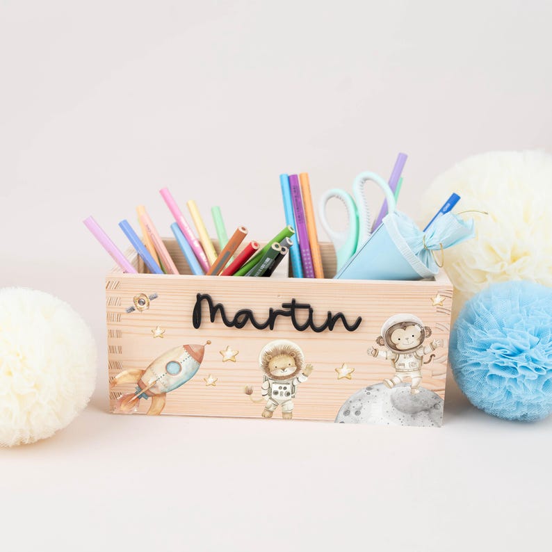 May include: A rectangular wooden pencil holder featuring the name Martin in black script. The front is decorated with illustrations of a rocket, a lion astronaut, and a monkey astronaut on the moon, filled with various coloured pencils and scissors.