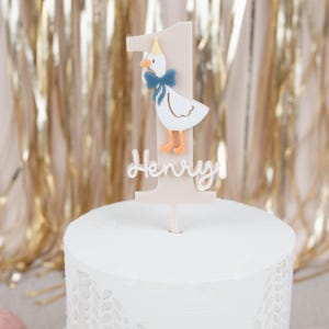 May include: A white cake with pink and white fondant icing. The cake is decorated with a white and orange goose topper with a blue bow and a pink and white number one. The topper also has the name "Henry" written on it.