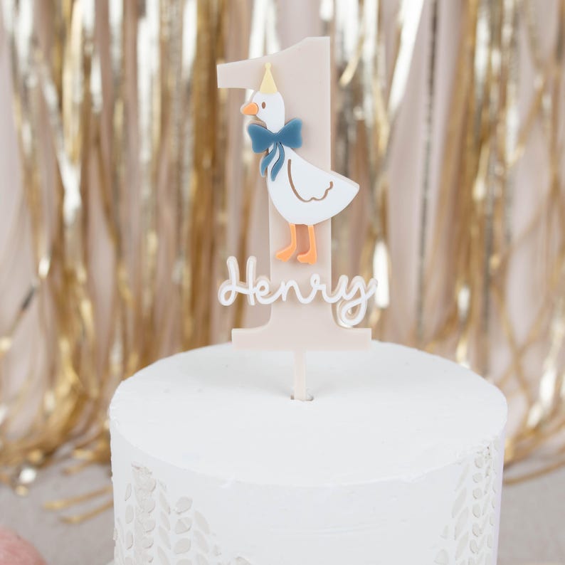 May include: A white cake with pink and white fondant icing. The cake is decorated with a white and orange goose topper with a blue bow and a pink and white number one. The topper also has the name "Henry" written on it.