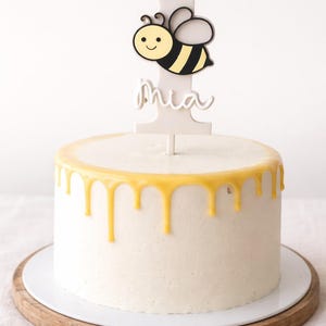 Op de afbeelding: Een ronde witte cake met gele glazuur die langs de zijkanten druipt, op een houten taartstandaard. Een witte taarttopper toont een cartoonbij en het nummer 1, met de naam "Mia" eronder.