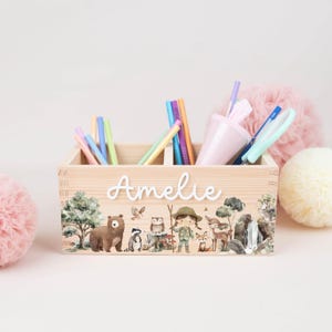 May include: A wooden desk organizer with the name "Amelie" in white script. The organizer is decorated with a woodland scene featuring a bear, owl, and a girl with a hat. Various pens and pencils are inside.