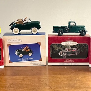 May include: Four Hallmark Keepsake ornaments of vintage vehicles: a yellow kiddie car, a green 1949 Graham Sport, a green 1937 Ford V-8 truck, and a red 1953 GMC truck. Each ornament sits atop its original box, featuring the vehicle's image and the Hallmark logo.