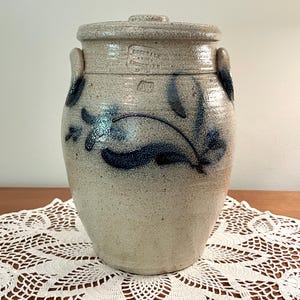 May include: A beige stoneware crock with a lid and two side handles, featuring a hand-painted dark blue floral vine design. The surface is speckled with a glossy finish. Stamped text reads Rockdale Union Stoneware and the initials AW are visible.
