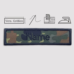 Könnte beinhalten: Ein Patch aus Tarnstoff mit schwarzen Nähten und dem Wort "Name" in schwarzem Faden gestickt. Der Patch kann auf Kleidung oder andere Gegenstände genäht oder gebügelt werden.