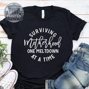 May include: A dark blue t-shirt with white text that reads "Surviving Motherhood One Meltdown At A Time".
