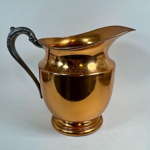May include: A metallic copper-colored pitcher with a rounded body, a wide pouring spout, and a dark, ornate, curved handle. The surface of the pitcher has a reflective, polished finish with subtle signs of wear and light oxidation near the handle.