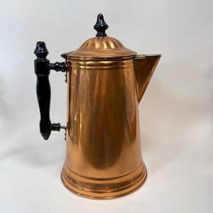 May include: A polished copper coffee pot featuring a tall, cylindrical body with a wide base. It includes a black handle attached to the side and a matching black finial on the domed lid. The spout is angular and extends from the upper rim.