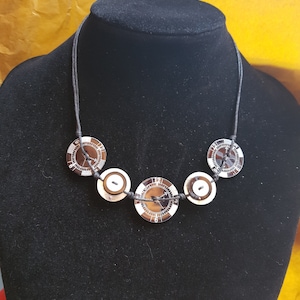 Può includere: Una collana con cordoncino nero con cinque bottoni argentati. I bottoni sono rotondi e hanno un design bianco e nero con numeri.