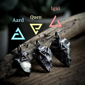 Könnte beinhalten: Drei silberne Pfeilspitzen-Anhänger mit verschiedenen Symbolen. Die Symbole sind Dreiecke mit einer Linie im Inneren. Die Symbole sind mit "Aard", "Quen" und "Igni" beschriftet.