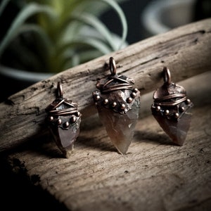 May include: Three arrowhead pendants with copper wire wrapping and copper beads. The arrowheads are a light brown colour and have a rough, natural texture.