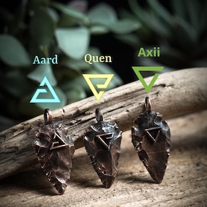 Può includere: Tre ciondoli a forma di punta di freccia in rame con diversi simboli. I simboli sono triangoli con linee all'interno. I simboli sono etichettati "Aard", "Quen" e "Axi" in testo verde e blu.
