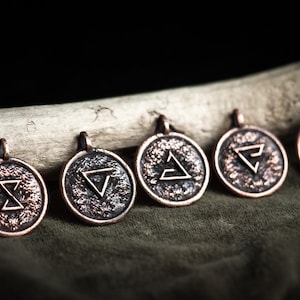 May include: Five bronze-colored pendant necklaces with different geometric symbols. The pendants are round with a textured surface and a loop at the top for a chain. They rest on a green fabric with a wooden branch in the background.