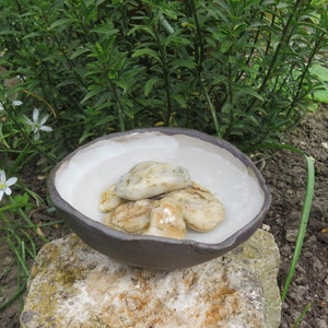 Könnte beinhalten: Eine kleine, dunkelgraue Keramikschale mit weißem Innenraum, gefüllt mit glatten, hellbraunen und weißen Steinen. Die Schale steht auf einem hellgrauen Stein.