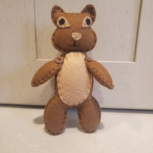 May include: A brown felt stuffed animal shaped like a bear with a tan felt belly and black eyes. The bear has two brown buttons on its chest.
