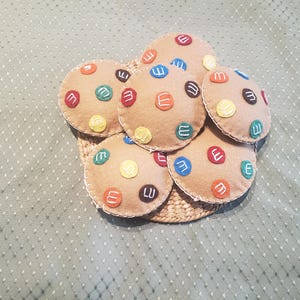 May include: A set of felt cookies with colorful M&M's on top. The cookies are arranged in a circle on a woven basket.