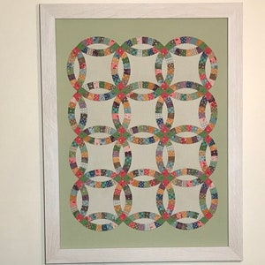 May include: A framed quilt with a green background and a pattern of interlocking rings in various shades of pink, red, blue, green, and yellow. The quilt is made of patchwork fabric and is displayed on a white wall.