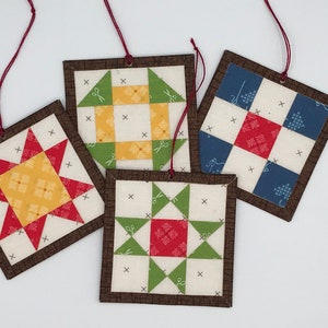 May include: Four handmade fabric ornaments with a brown border. Each ornament features a different quilt block pattern in red, yellow, green, and blue fabrics. The ornaments have a red string attached for hanging.