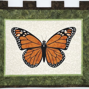 May include: A brown and black butterfly with white spots is framed in a green and brown fabric. The butterfly is centered on a white background with a green and brown border.