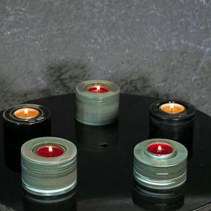 Puede incluir: Cinco candelabros para velas de té con velas encendidas. Los candelabros son cilíndricos, con una hendidura circular para sostener las velas. Las velas son rojas y naranjas, y los candelabros son negros y verdes.