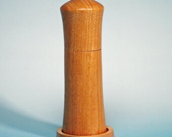 Könnte beinhalten: Eine Pfeffermühle aus hellem Holz. Die Mühle hat einen zylindrischen Körper mit ausgestelltem Boden und abgerundeter Oberseite. Sie steht auf einem kleinen, kreisförmigen Holzsockel. Die Holzmaserung ist sichtbar. Etwa 18 cm hoch.