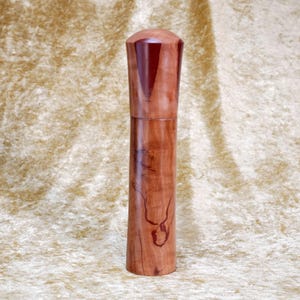 Könnte beinhalten: Eine zylindrische Pfeffermühle aus Holz mit glatter, polierter Oberfläche und sichtbarer natürlicher Holzmaserung in hellbraunen und beigen Farbtönen, gekrönt von einem dunkelrotbraunen Akzentstück an der abgerundeten Kappe.