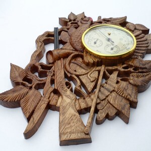 May include: A wooden wall hanging with a carved design of a hunting scene. The scene includes two rifles, a hunting horn, and leaves. A round barometer with a gold rim is in the centre of the design.