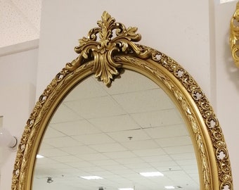 Puede incluir: Un espejo grande de forma ovalada con un marco dorado y ornamentado. El marco tiene un diseño detallado con tallas intrincadas y una pieza central elevada.