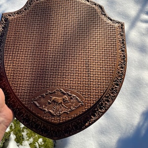 May include: A brown wooden plaque with a woven pattern and a carved image of a dog running through tall grass. The plaque has a decorative border.