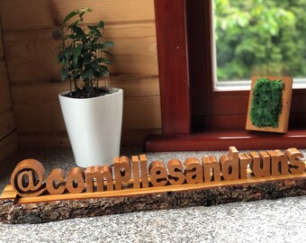 May include: Wooden sign with the text "@compilesanddrums" on a natural bark base. The sign is sitting on a tiled surface.