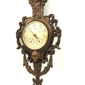 May include: A vintage, ornate, dark brown, cast iron barometer with a gold face and three dials. The barometer has a decorative design with a face and a floral motif.