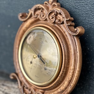 May include: A vintage-style, round barometer with a gold-coloured face and a decorative, ornate brown frame. The barometer features a dial with various weather indicators and a needle pointing to the current atmospheric pressure. The frame has detailed scrollwork.