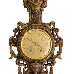 May include: An antique-style barometer with a bronze finish. The circular gauge has a gold-coloured face with multiple dials and a needle indicating atmospheric pressure. The decorative frame includes scrollwork, a crest, and a carved face at the bottom.