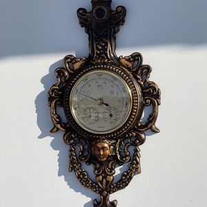 May include: An antique-style, ornate, gold-toned barometer with a black face and a decorative, black and gold frame. The barometer has three dials: one for temperature, one for humidity, and one for barometric pressure. The frame is decorated with a cherub's face and floral motifs.