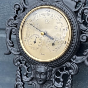May include: An ornate black metal weather station with a gold-coloured face. The face displays a barometer, thermometer, and hygrometer. The design includes decorative scrollwork and a sculpted face. The barometer reads in mmHg.