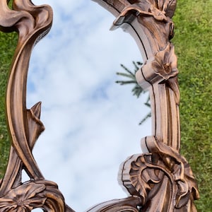 May include: Ornate, Art Nouveau-style mirror with a bronze-colored, carved wooden frame. The frame features flowing lines, floral and leaf motifs, and an oval-shaped mirror reflecting a cloudy sky. Decorative wall art.