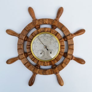 Puede incluir: Un barómetro de madera en forma de rueda de barco con un borde dorado y una cara blanca. El barómetro tiene tres diales: uno grande para la presión barométrica, uno más pequeño para la temperatura y un tercero para la humedad. La rueda tiene ocho radios y está hecha de madera oscura.