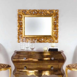 May include: A gold-framed mirror with ornate detailing hangs above a gold-toned dresser. The dresser has two drawers with crystal knobs. Clear glass vases and decorative spheres sit on top of the dresser, creating a luxurious aesthetic.