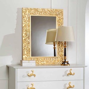 May include: A gold-framed mirror with ornate carvings is displayed above a white dresser. A matching gold and black lamp with a beige lampshade sits to the right of the mirror. The dresser has gold-colored handles.